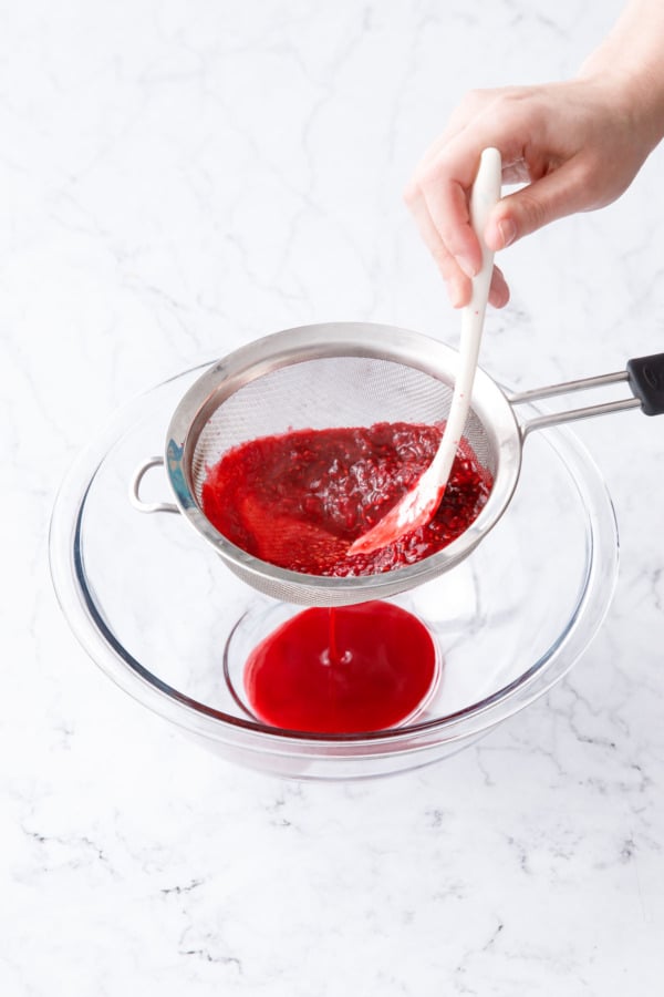 Straining raspberry mixture through a fine mesh sieve held over a glass mixing bowl.