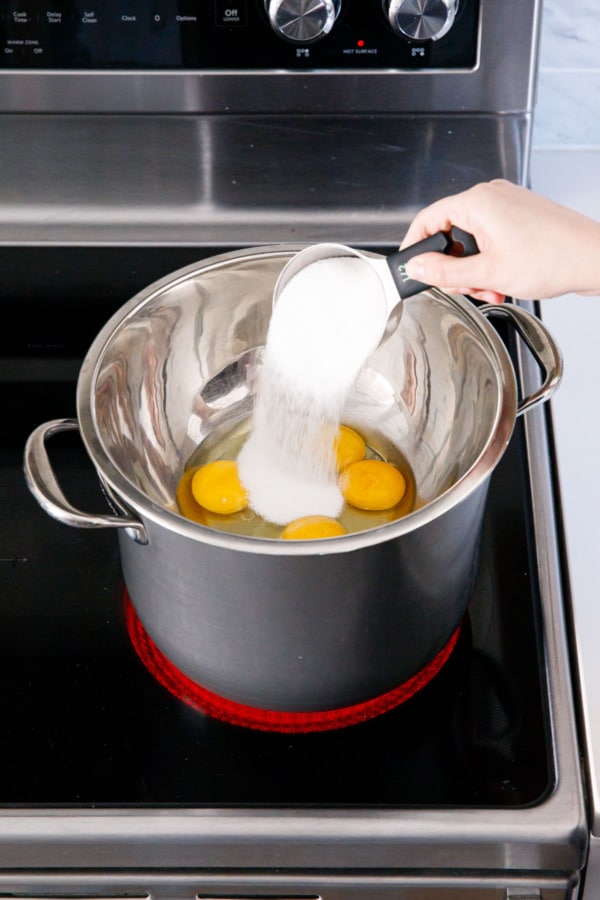 Pouring sugar into metal mixing bowl made into a double boiler set over a saucepan on a stovetop.