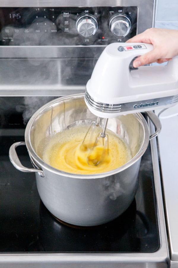 Hand mixer whipping eggs and sugar in a mixing bowl set over a saucepan of simmering water.