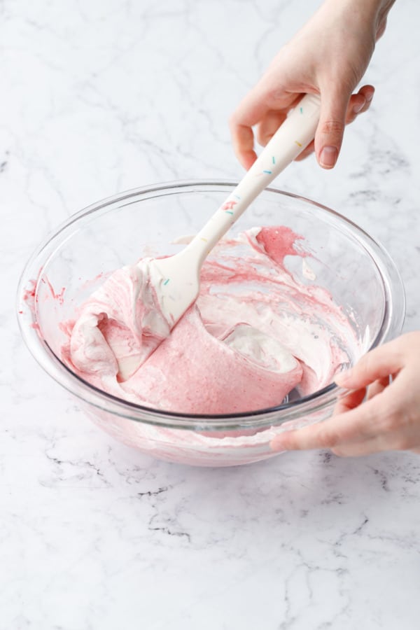Folding whipped cream into raspberry base mixture in a glass mixing bowl with large silicone spatula.
