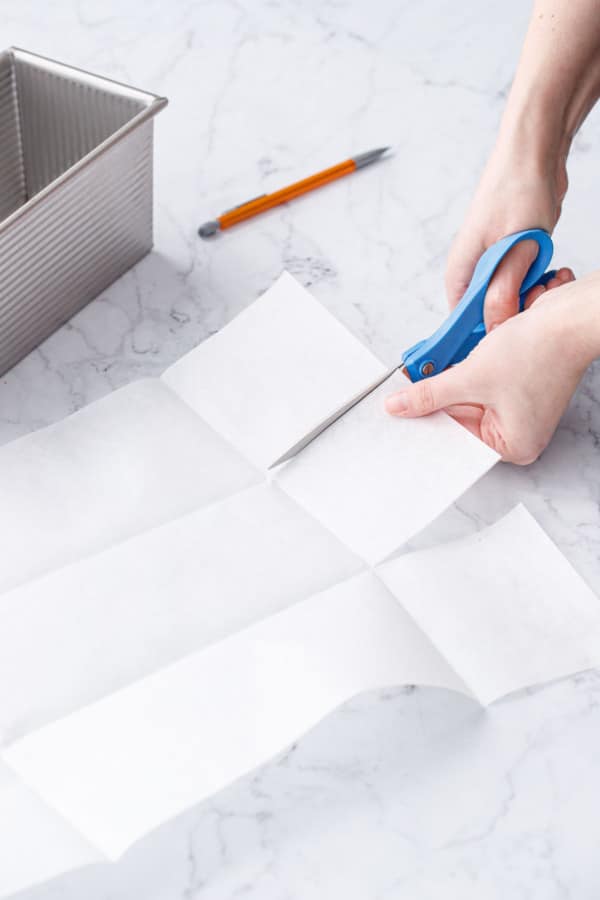 Cutting a folded piece of parchment paper to line a square loaf pan.