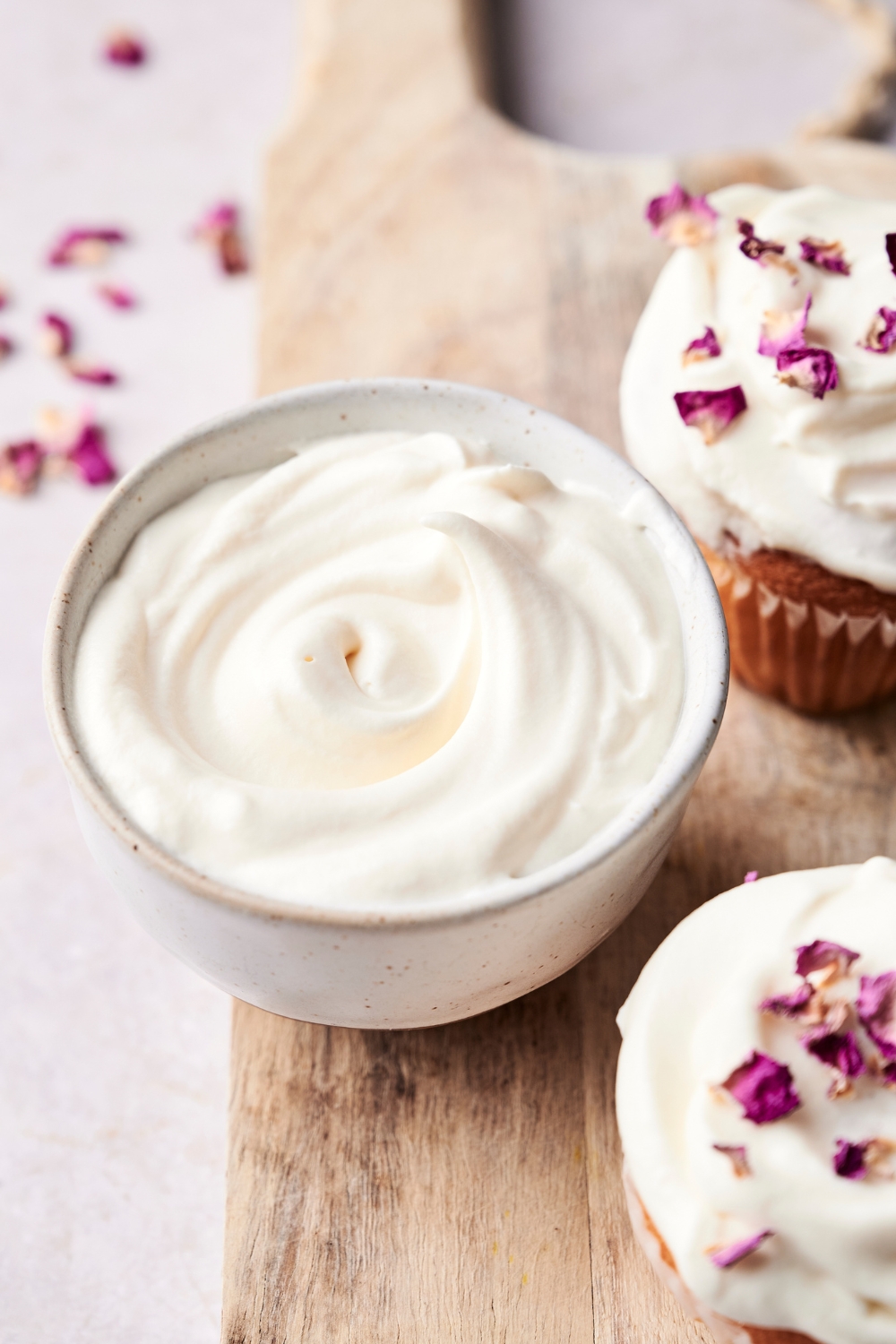 A small bowl with creme fraiche frosting sitting on a wooden board next to two cupcakes.