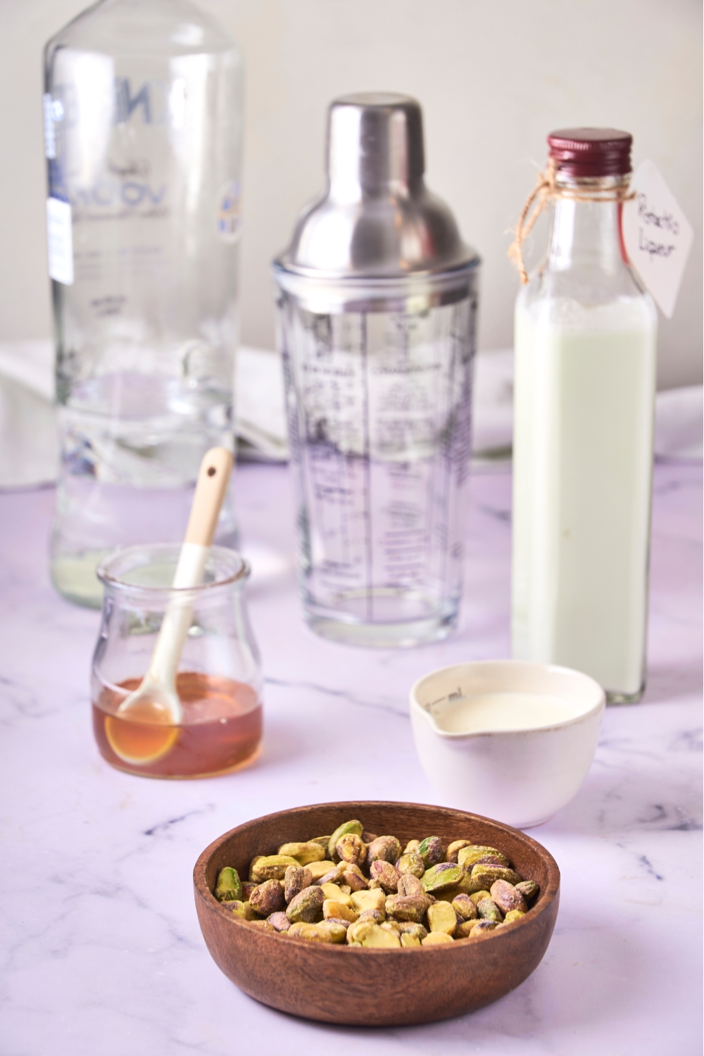 A countertop with multiple bowls and containers with ingredients like pistachios, vanilla vodka, pistachio liqueur, and honey.