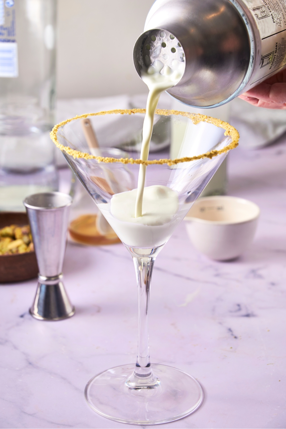 A martini glassed rimmed with crushed pistachios being filled by the cocktail shaker.