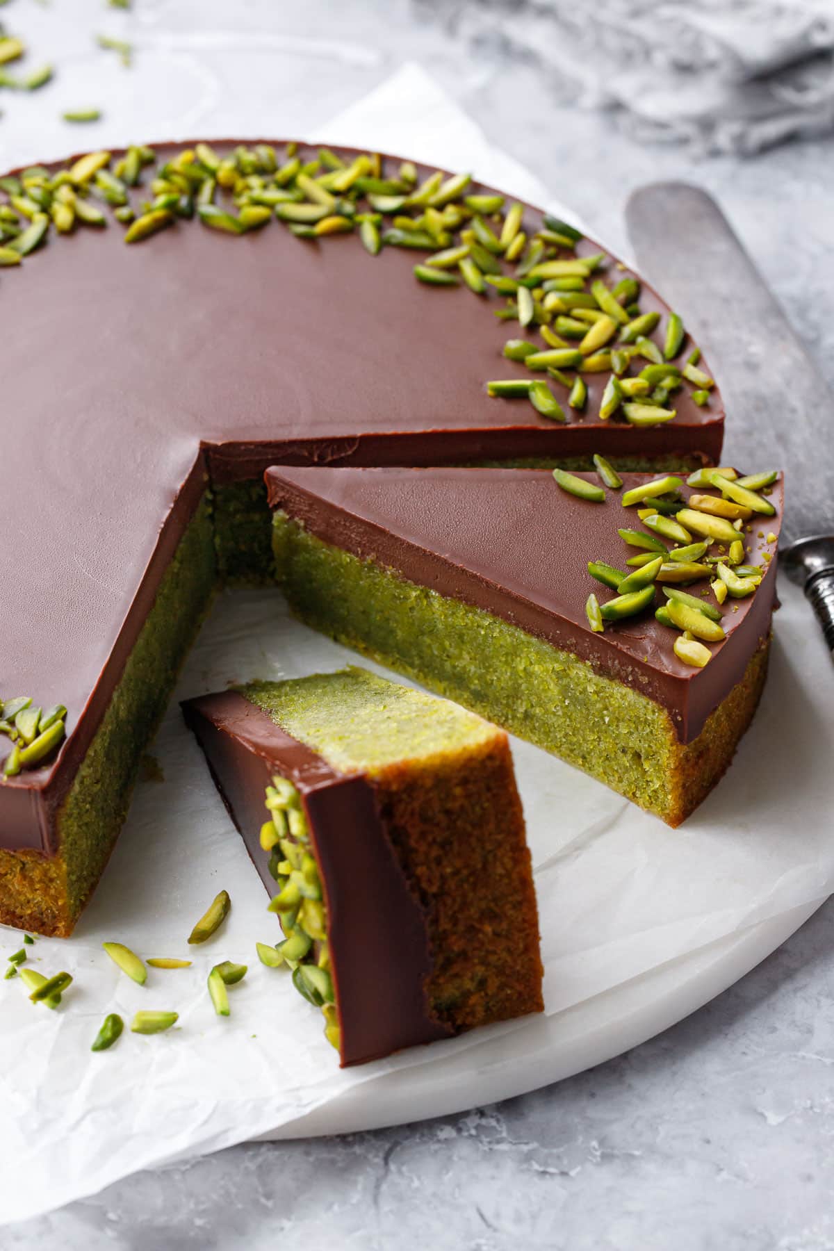 Flourless Pistachio Cake on a flat cake plate with two slices cut out, one on its side with slivered pistachios falling off.