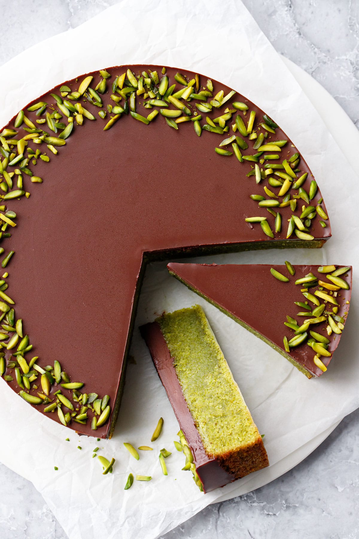 Overhead, two cut slices of Chocolate Ganache-topped Flourless Pistachio Cake, one on its side showing the bright green interior, with a border of slivered pistachios.