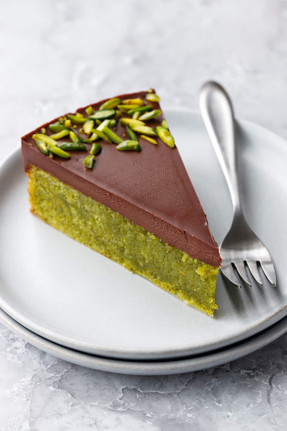 Closeup, cleanly cut slice of Flourless Pistachio Cake topped with Chocolate Ganache and slivered pistachios, on a stack of gray plates with a silver fork.