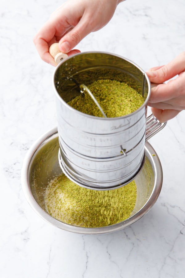 Sifting pistachio and almond flours together with a metal sifter into a silver stainless bowl.