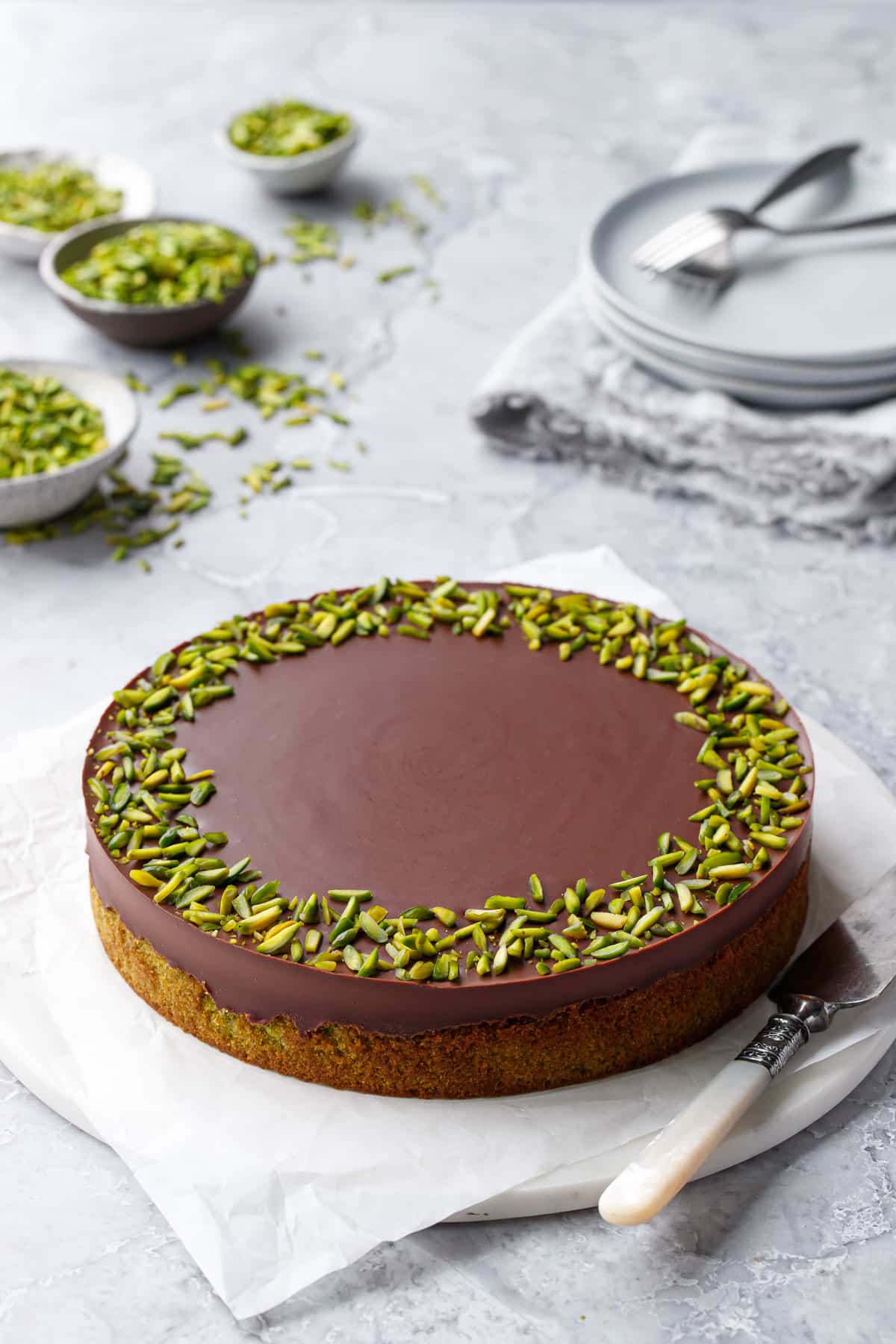 Flourless Pistachio Cake topped with a smooth layer of Chocolate Ganache and a border of bright green slivered pistachios, on a plate with cake server, bowls of pistachios in the background.