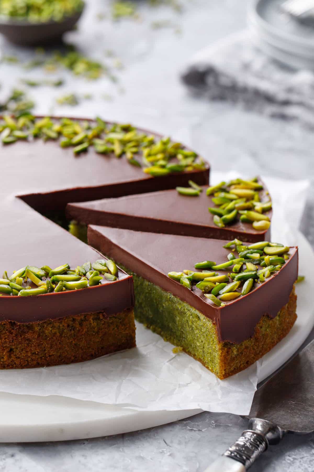Two cut slices of Flourless Pistachio Cake with Chocolate Ganache and a border of bright green slivered pistachios, on a piece of crinkled parchment paper sitting on a marble cake plate.