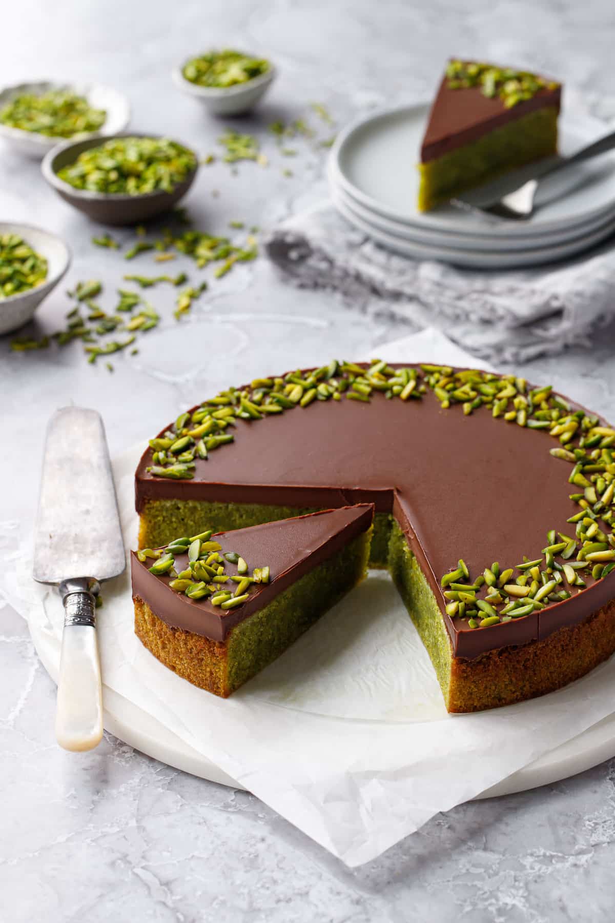 Flourless Pistachio Cake with Chocolate Ganache and topped with slivered pistachios, on a gray background with cake server, cut slice and more pistachios in the background.