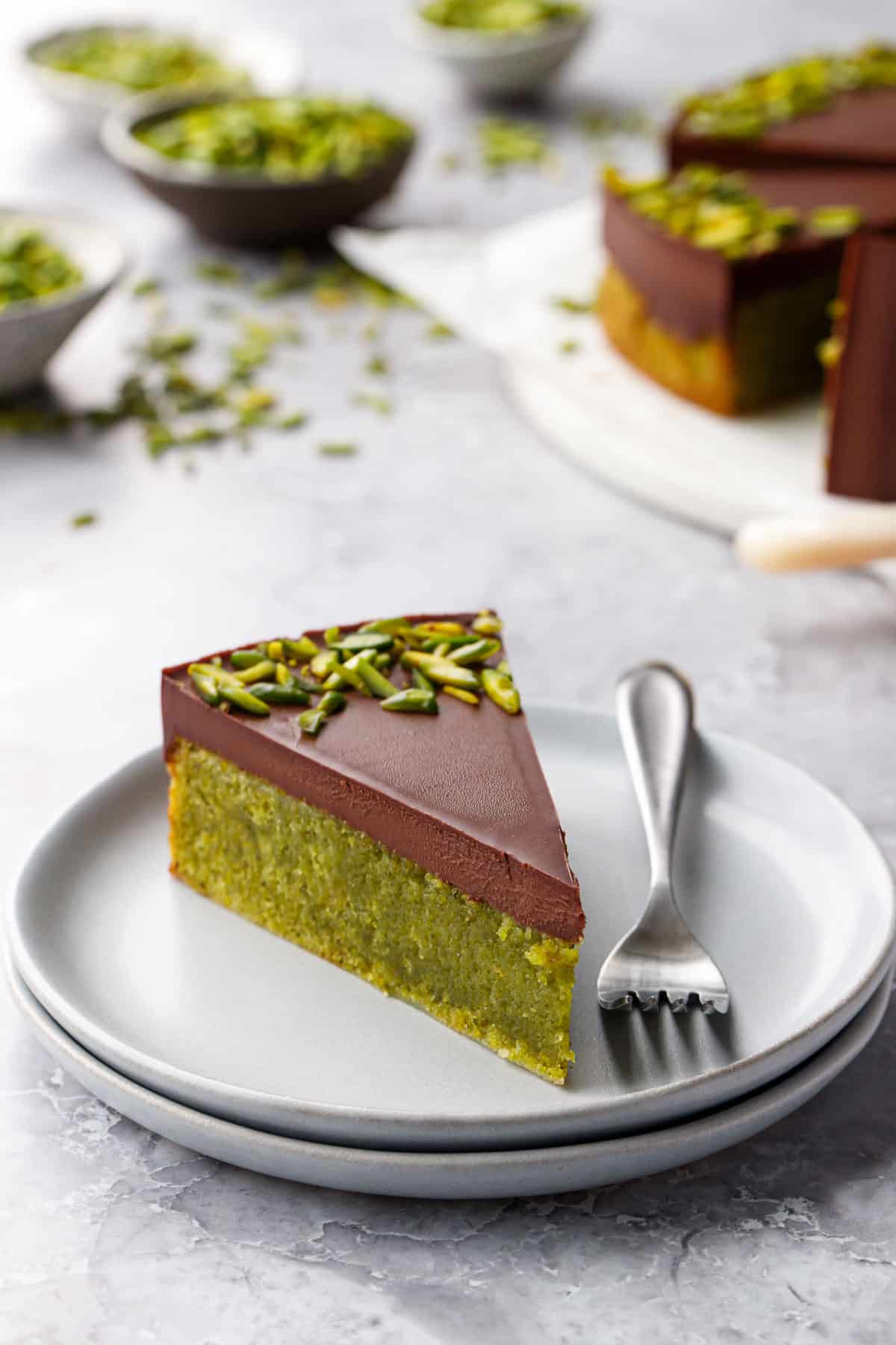Closeup clean cut slice of bright green Flourless Pistachio Cake, topped with Chocolate Ganache and green slivered pistachios on a gray plate with fork.