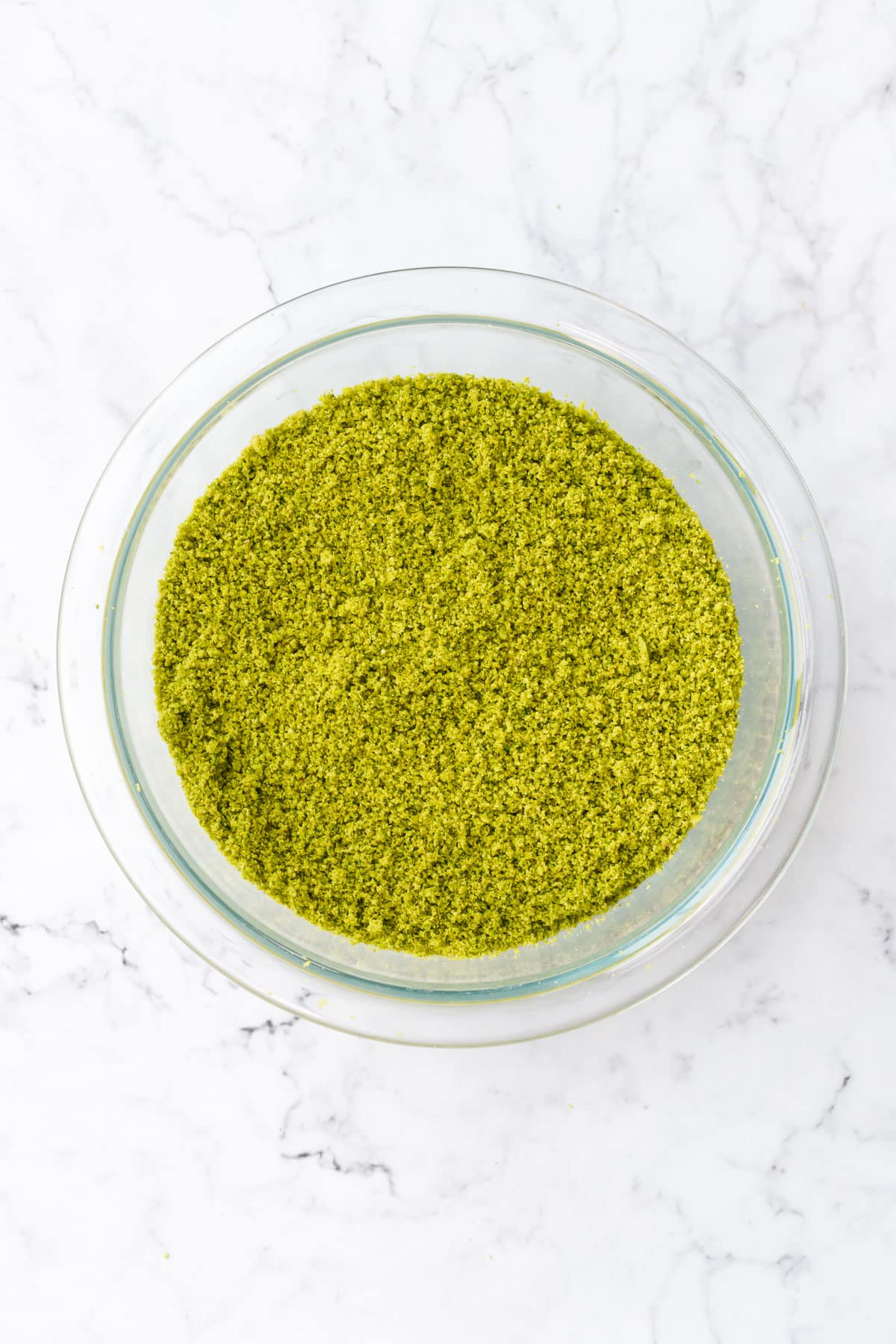 Overhead, bright green Homemade Pistachio Flour in a glass bowl on a marble background.