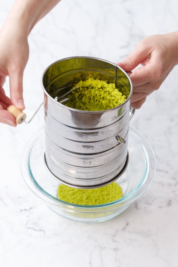 Sifting homemade pistachio flour in a stainless steel sifter set over a glass mixing bowl.