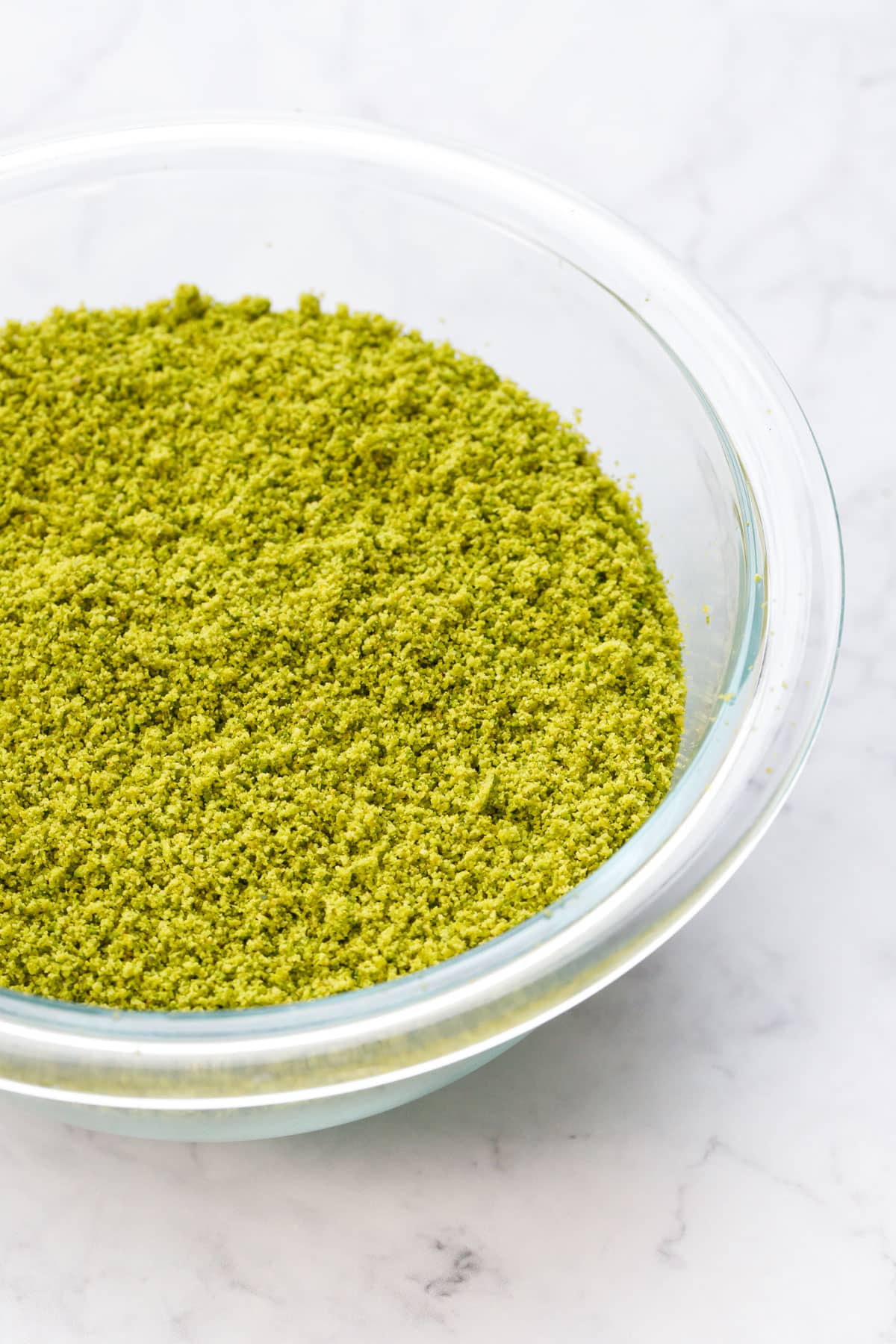Bright green homemade pistachio flour in a glass mixing bowl on a marble background.