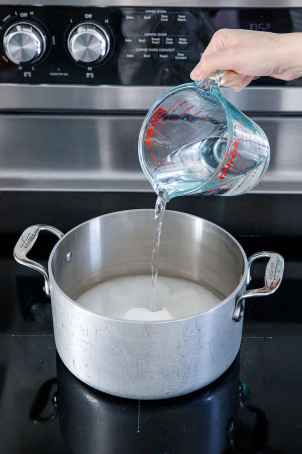 Pouring water over granulated sugar in a 4qt stainless saucepan on the stovetop.