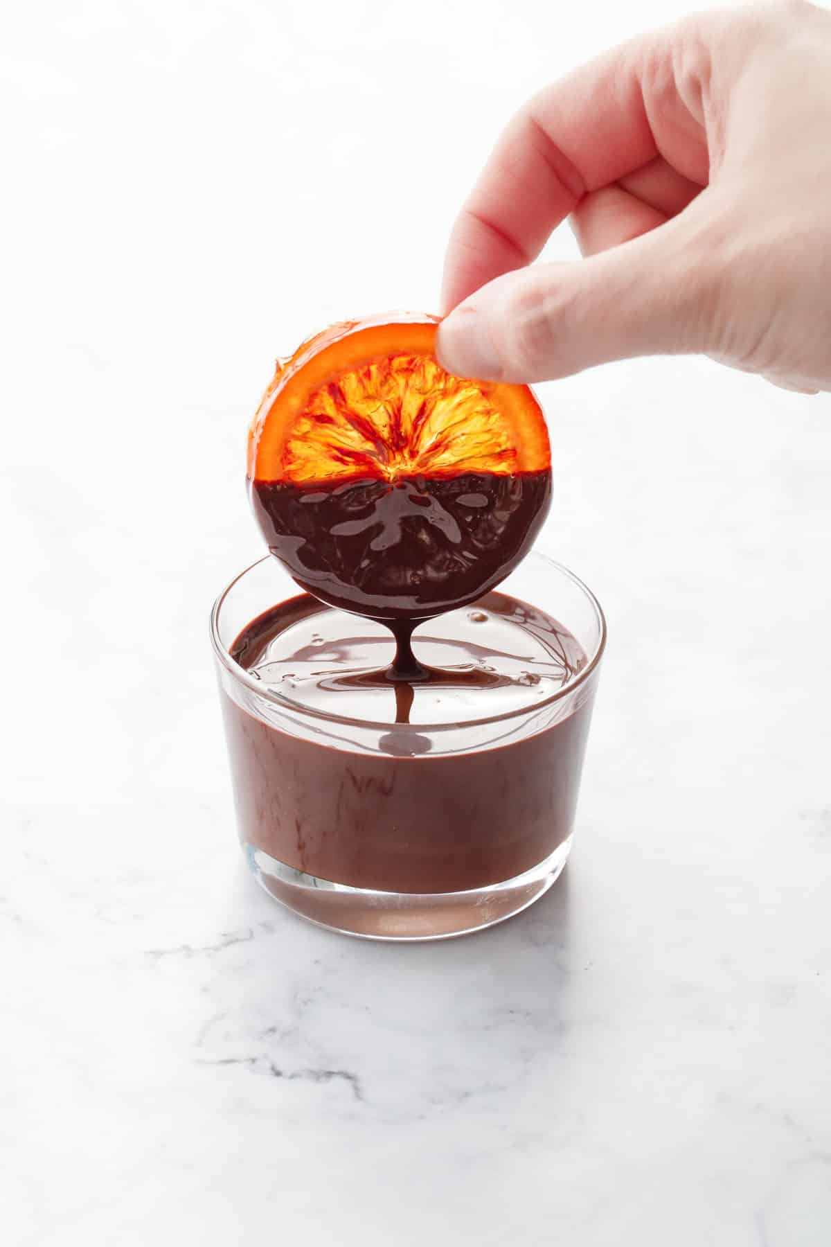 Hand dipping a glass-like transparent slice of candied blood orange into a small glass of melted dark chocolate.