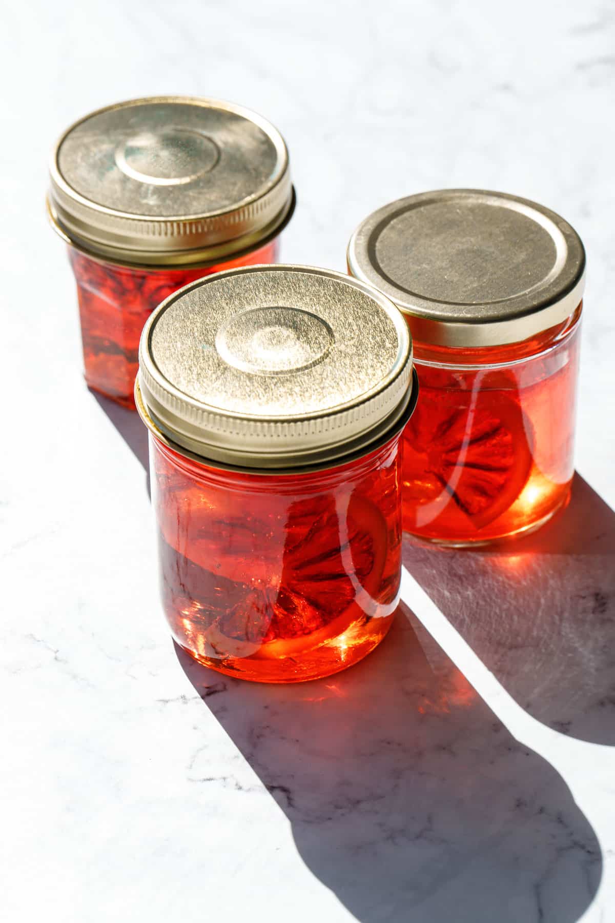 Jars of red candied blood orange syrup in glass jars with gold lids, backlit to show the slices of candied orange floating inside.