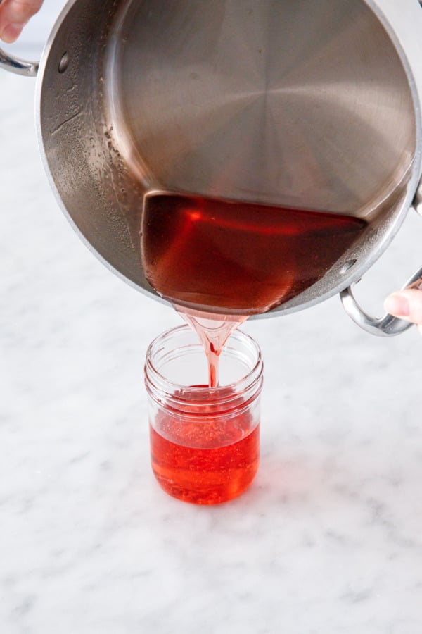 Pouring bright orange-red syrup from saucepan into a glass jar.