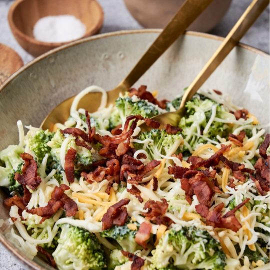 A large serving bowl holds Chicken Salad Chick Broccoli Salad topped with bacon and shredded cheese. There is a pair of golden serving utensils in the bowl, as well.