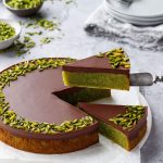 Cake server lifting a cut slice of Flourless Pistachio Cake with a bright green interior and a layer of chocolate ganache on top.