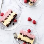 Overhead, marble background and three glass plates with cross-section slices of Raspberry & Passionfruit Semifreddo with Chocolate Crumbs, and a few frozen raspberries scattered around.