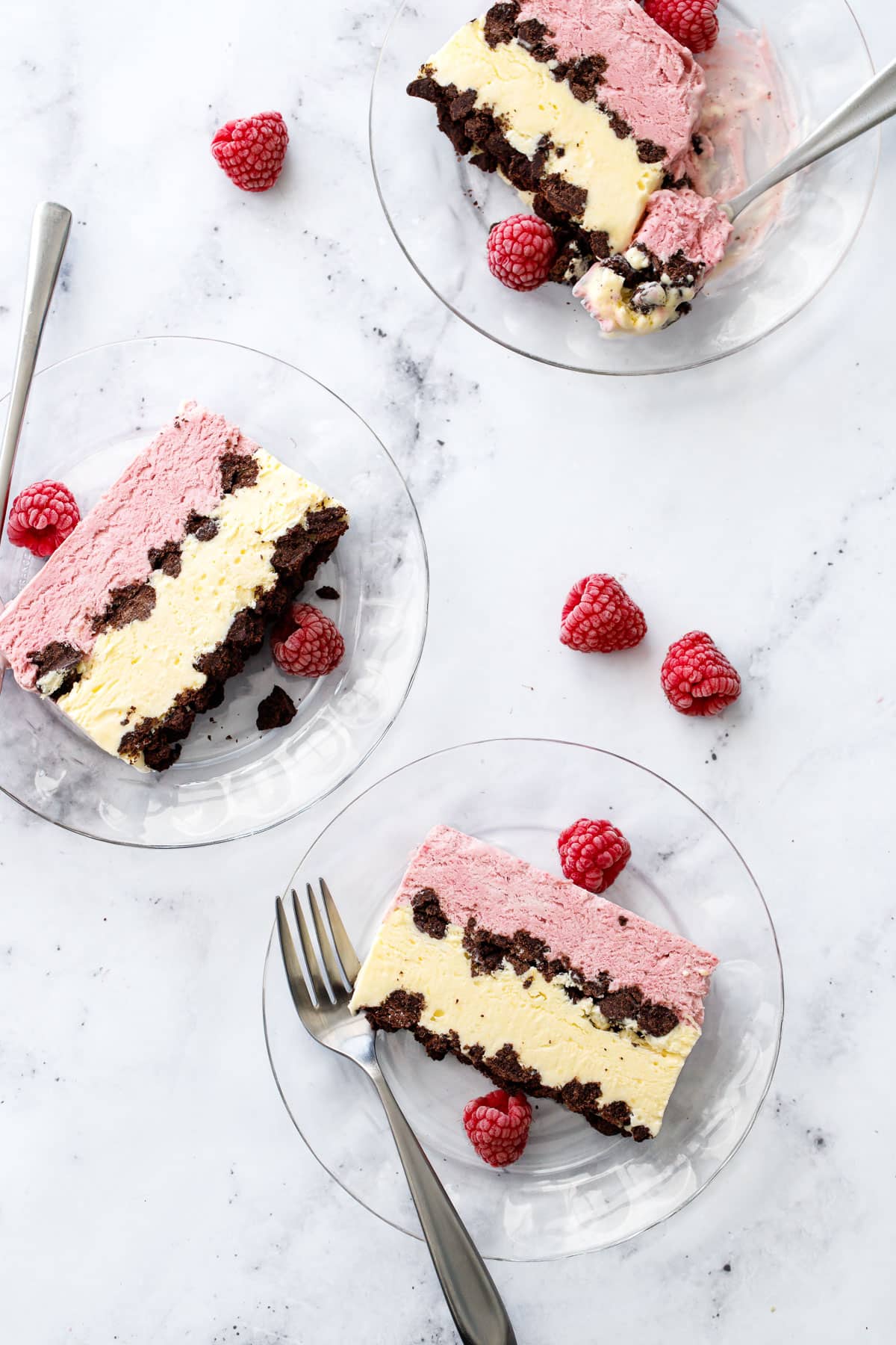 Overhead, marble background and three glass plates with cross-section slices of Raspberry & Passionfruit Semifreddo with Chocolate Crumbs, and a few frozen raspberries scattered around.