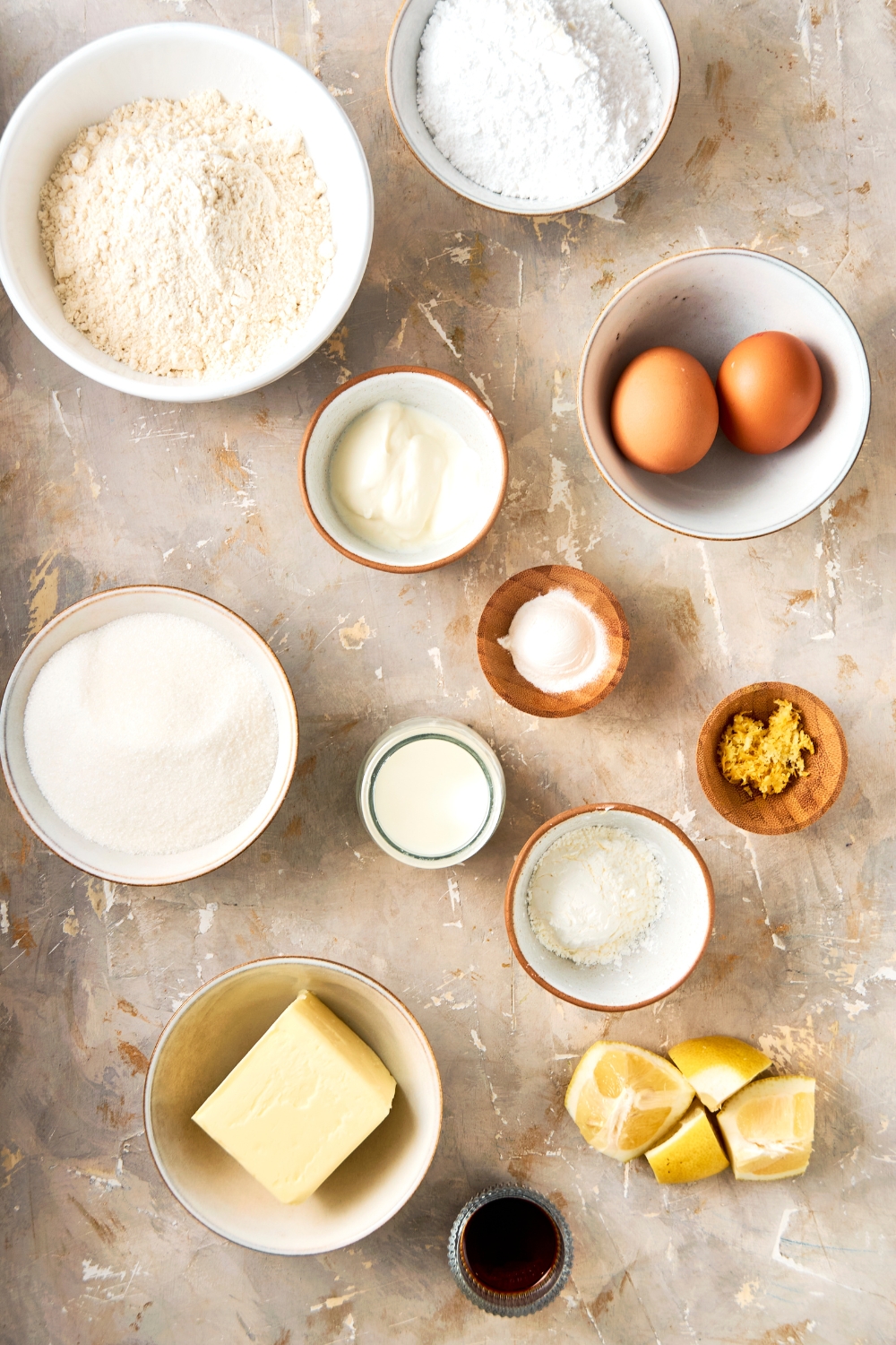 A countertop with multiple bowls with ingredients like flour, sugar, butter, greek yogurt, eggs, lemon zest, baking powder, salt, milk, vanilla, and sliced lemons.