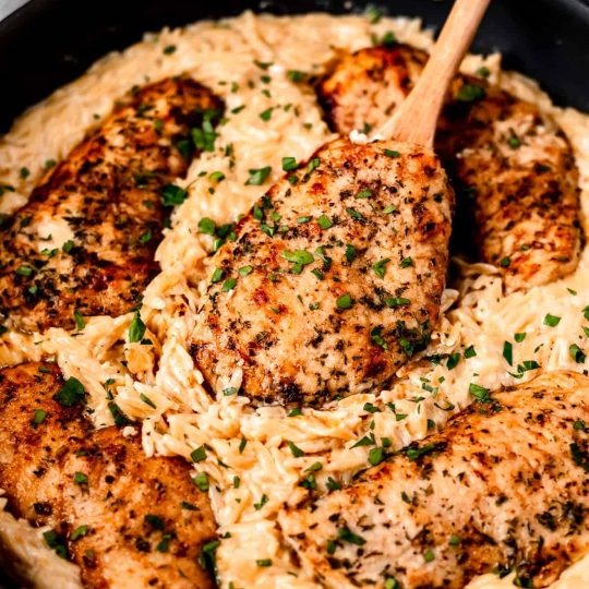 Parmesan Chicken Orzo in a skillet with a wood spoon lifting up a piece of chicken.
