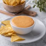 Taco Bell nacho cheese sauce in a white serving bowl atop a white plate filled with tortilla chips.