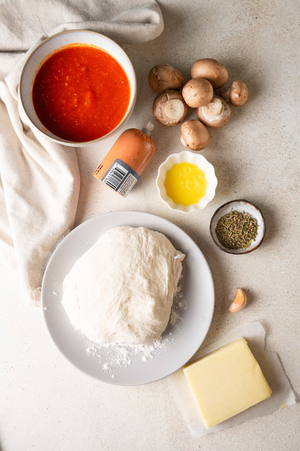Pizza dough, butter, mozzarella cheese, seasonings, pepperoni, mushrooms, and crushed tomatoes are all in separate containers on a counter top.