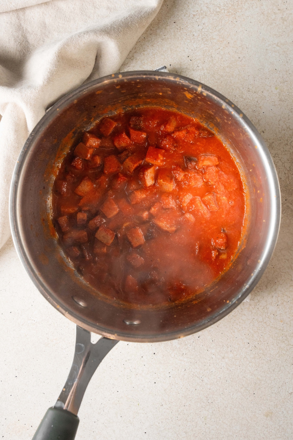 A silver saucepan holds crushed tomatoes and pepperoni slices.
