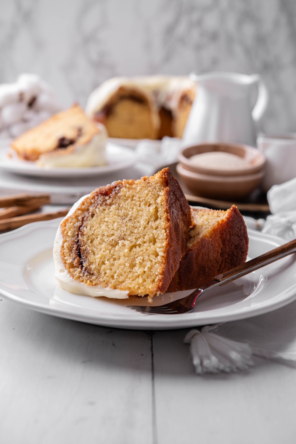 Two pieces of cinnamon bundt cake are on a white plate. There is a fork on the plate. Another piece of cake is on a plate in the background.