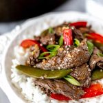Close up of ginger beef stir fry with sugar snap peas and red peppers in a white bowl with rice with a skillet partially showing in the background.