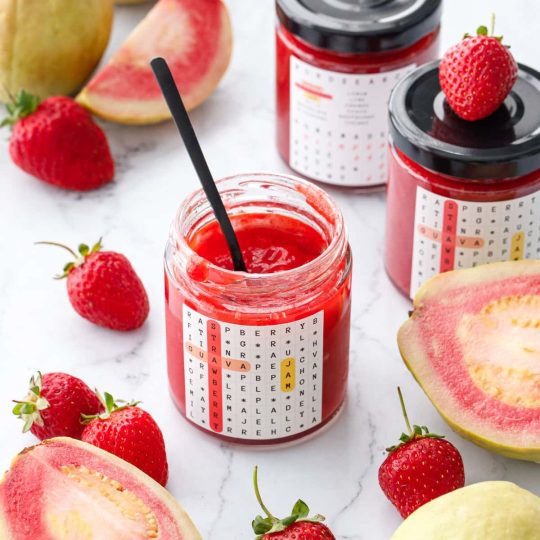 Jars of Guava Strawberry Jam on a marble background with fresh strawberries and whole and cut pink guavas scattered around.