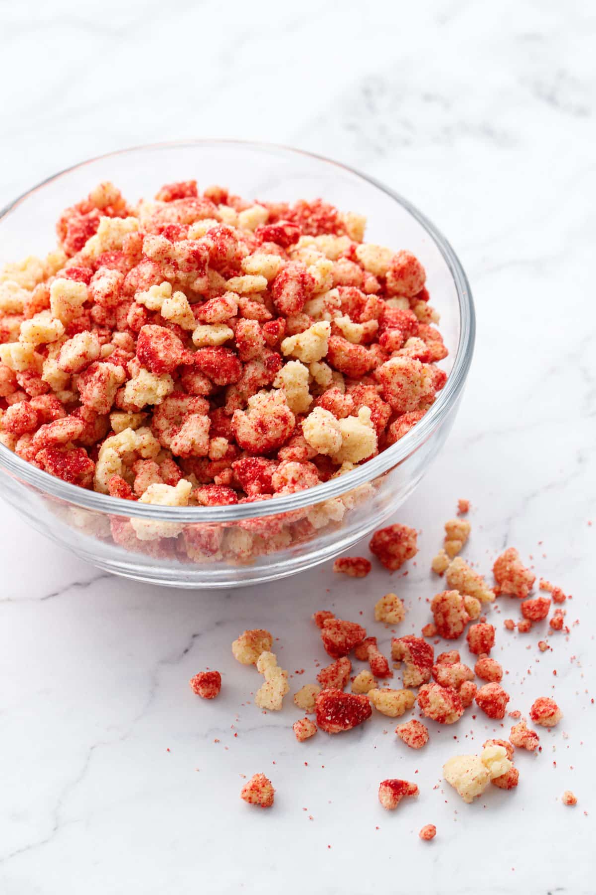 Glass bowl of Strawberry Shortcake Crunch Topping on a marble background, with a few crumbs scattered alongside the bowl.