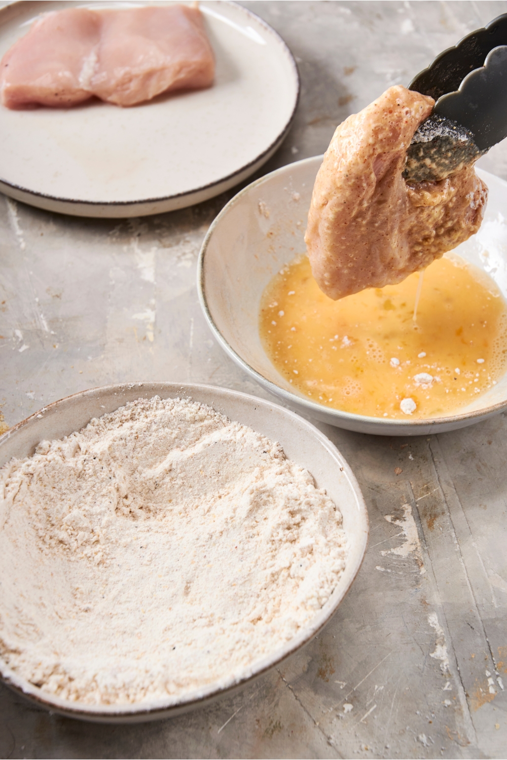 Someone dredges a chicken breast in egg wash. A shallow bowl of flour mixture is nearby.
