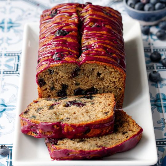 Loaf of Blueberry Banana Bread on a white rectangular plate with two slices lying down on a blue and white tile background.