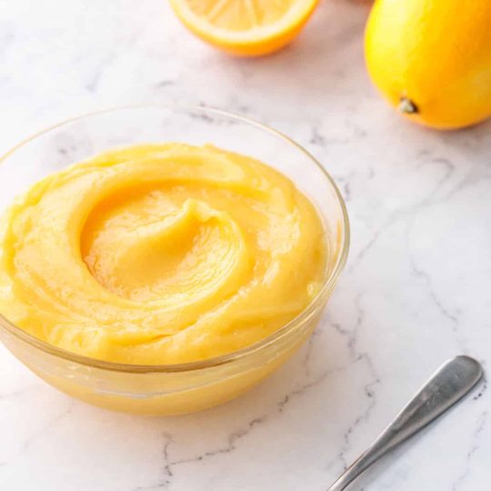 Glass bowl filled with Meyer Lemon Curd on a marble surface, with a spoonful of lemon curd in the foreground and a few fresh Meyer lemons out of focus in the background.