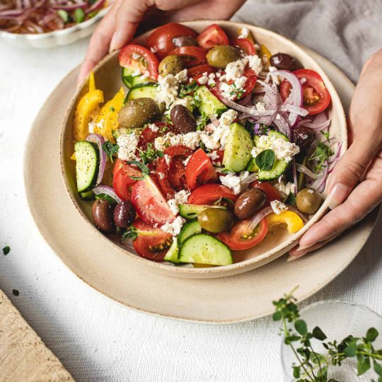 Big colorful bowl of Simple Homemade Greek Salad with hands placing it on table